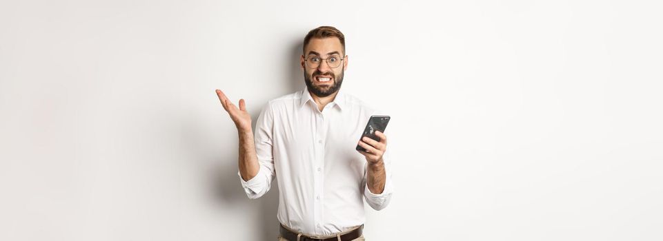 Confused businessman reading strange message on mobile phone, looking annoyed, standing over white background