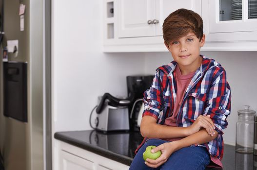 Apples are yum. a young boy sitting on the kitchen counter with an apple in his hand.