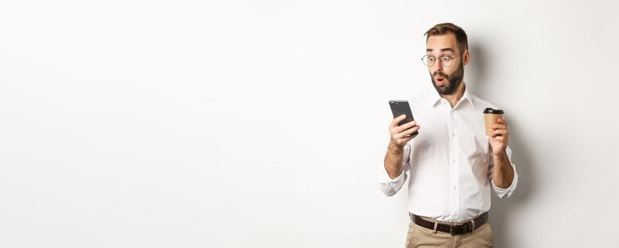 Businessman drinking coffee and looking surprised at message on mobile phone, standing amazed over white background.