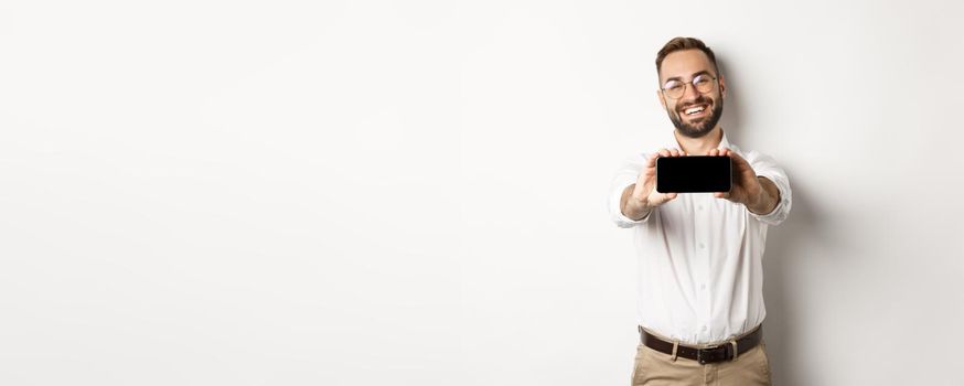Happy business man showing mobile screen, holding phone horizontally, standing satisfied against white background.