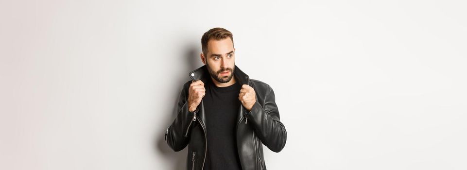 Attractive bearded man in leather biker jacket looking aside, standing confident against white background