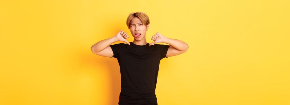 Portrait of sassy asian handsome guy showing thumbs-down and stick tongue disappointed, judge something bad, yellow background