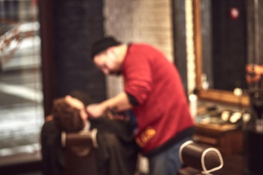 A strong handsome man getting shaved by a professional barber in a barbershop. Blurred image.