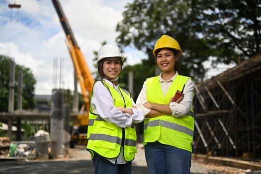 Architect women in safety helmet and reflective jackets standing with arms crossed font of industrial building construction site