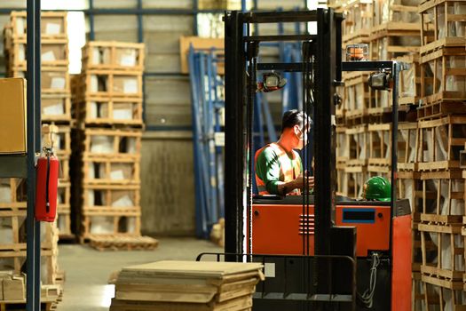 Warehouse worker in uniform loading cardboard boxes with forklift in warehouse full of shelves with goods