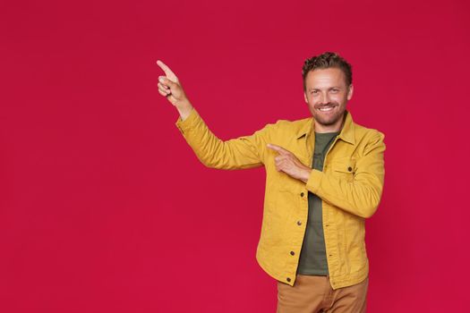 Happy bearded man in yellow jeans jacket pointing sideways up introducing copy space, mock up advertisement isolated on red background. Charming smiling freelancer introduce your business offer