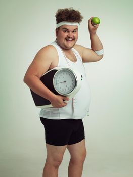 Its the fit life for me. Portrait of an overweight man holding a apple and a scale.