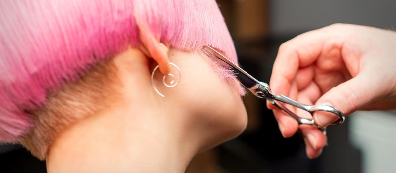 Cutting female hair. Hand of a hairdresser cutting short pink hair of young white woman at the hair salon