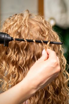 Hands of hairstylist curl wavy hair of young woman using a curling iron for hair curls in the beauty salon rear view.