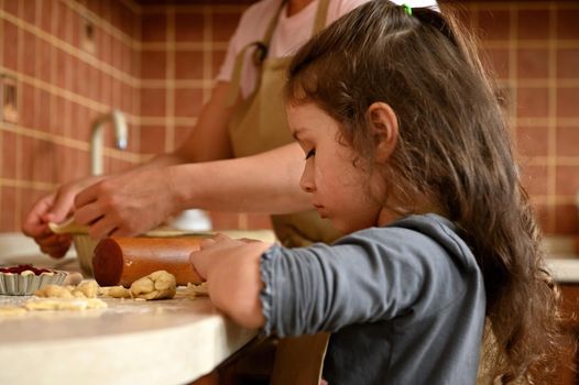Beautiful European little girl with long hair, using a rolling pin, rolling out the dough, cooking a pie with her mother