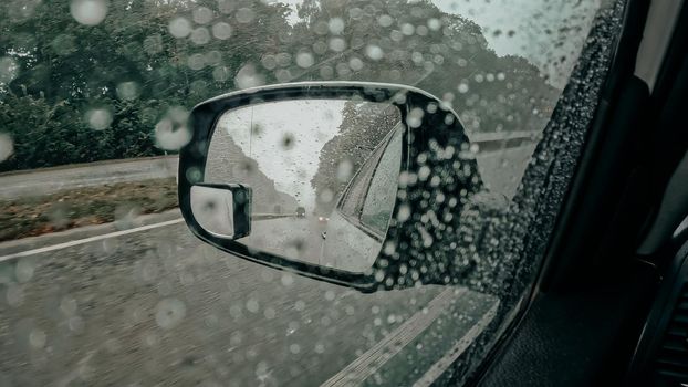 Side drivers car mirror with rain droplets. Driving automobile while rainy weather, busy road