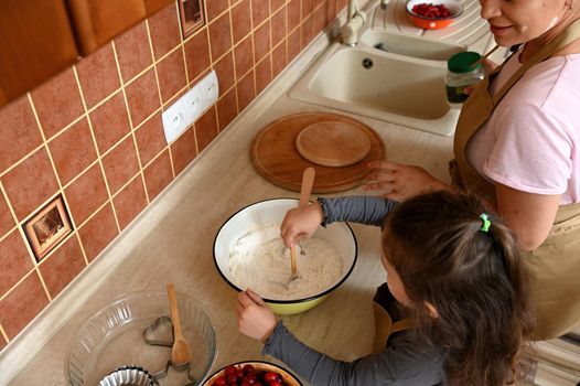 View from above to a little girl kneading dough for cherry pie, helping her mother in the kitchen. Child learns culinary