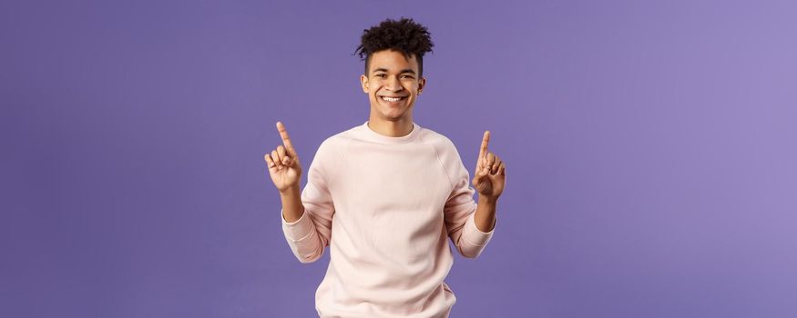 Waist-up portrait of joyful good-looking hispanic man with dreads, promoting product or company banner hanging on top, pointing fingers up, smiling satisfied, recommend subscribe or click link