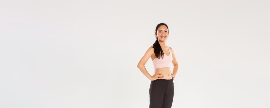 Full length of sweaty smiling fitness girl, female athelte in active wear looking pleased while workout, training in gym, satisfied with exercises, coach helping gain perfect body, white background
