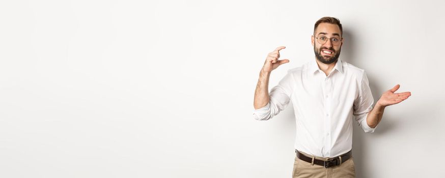 Businessman showing small gesture, looking displeased, shrugging confused, standing against white background