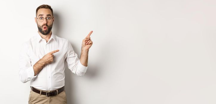 Impressed businessman showing interesting promo offer, pointing fingers right, standing amazed against white background