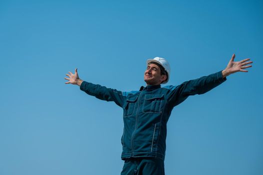 A caucasian man in work clothes and a construction helmet stands against the background of a blue sky with his arms outstretched to the sides.