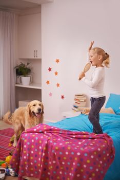 A star in the making. A little girl jumping on her bed and singing into her hairbrush while her dog watches.