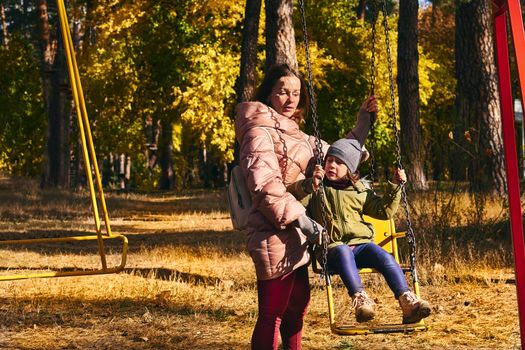 Young cute mother shakes her child daughter on a swing in a bright autumn park