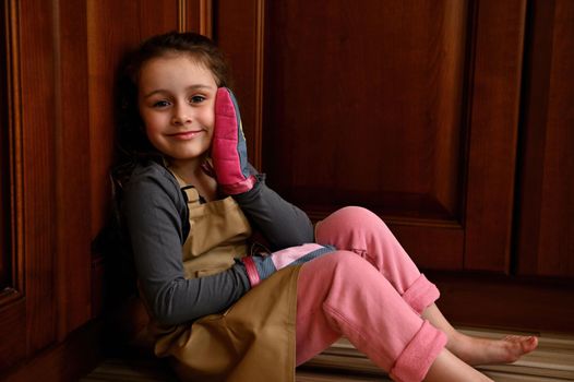 Portrait of a pretty adorable child girl, little baker confectioner in chef apron and kitchen mittens, smiling to camera