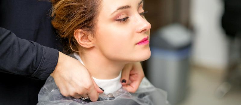 A hairdresser is covering a female neck with a cape for a female client in a beauty salon, close up.