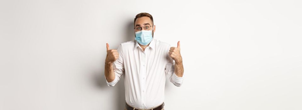 Covid-19, social distancing and quarantine concept. Impressed man in face mask showing thumbs-up and shrugging, standing amazed against white background