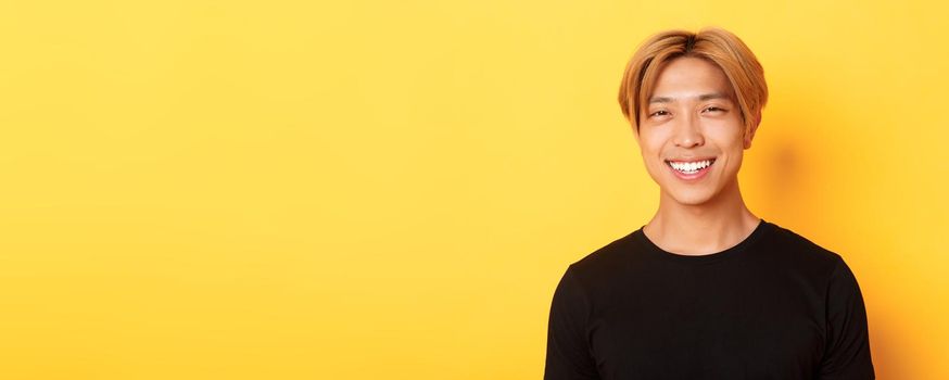 Close-up of handsome stylish asian guy with fair hair, smiling happy over yellow background