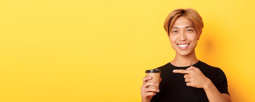 Close-up of satisfied handsome asian blond guy, smiling delighted and pointing finger at delicious cup of coffee, standing over yellow background