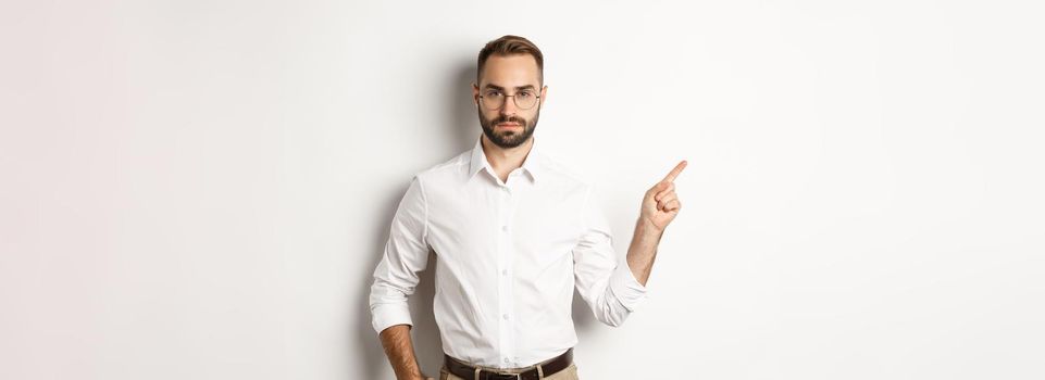 Serious bearded man pointing finger left, showing advertisement, standing over white background