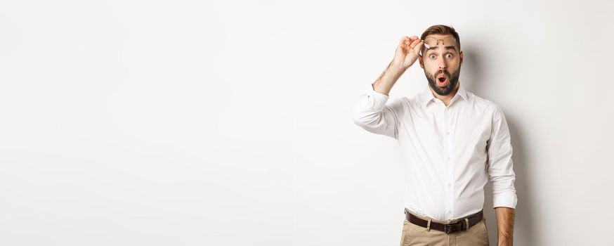Surprised businessman taking-off his glasses, looking with amazement at camera, standing over white background