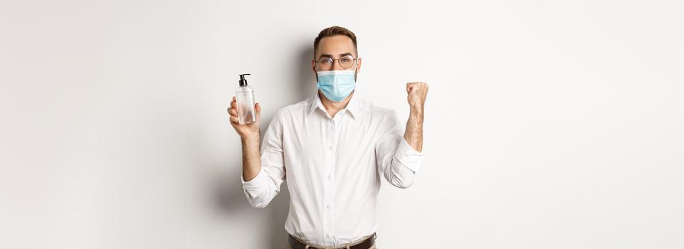 Covid-19, social distancing and quarantine concept. Cheerful manager in medical mask showing hand sanitizer, standing over white background