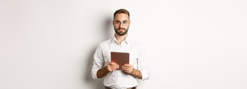 Serious employer working with digital tablet, reading in glasses, standing over white background