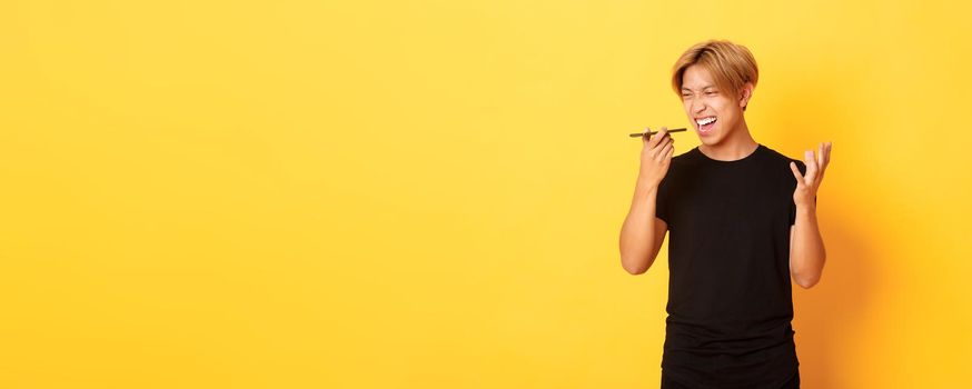 Portrait of angry asian man yelling mad at mobile phone speaker, record furious voice message, standing yellow background