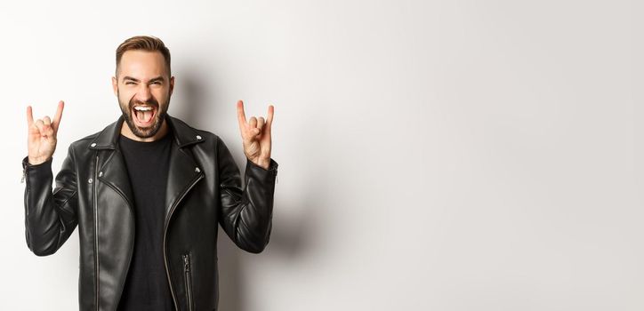 Cool adult man in black leather jacket, showing rock on gesture and tongue, enjoying music festival, standing over white background