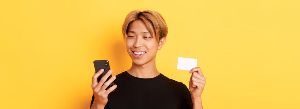 Close-up of handsome stylish asian guy shopping online, looking at mobile phone and smiling, showing credit card, standing over yellow background