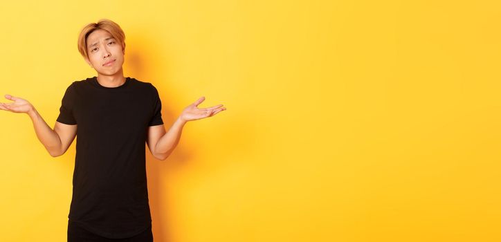 Portrait of clueless asian blond guy, wearing black clothes, shrugging and looking puzzled, standing yellow background