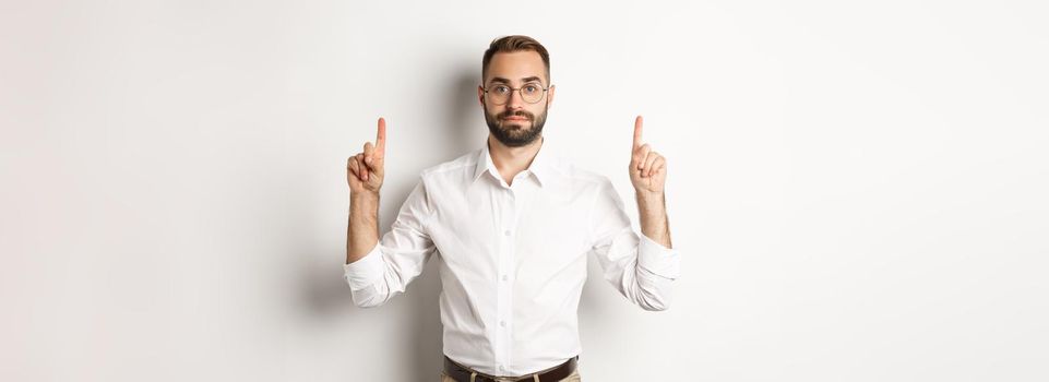 Young bearded businessman pointing fingers up, showing promo offer, standing over white background