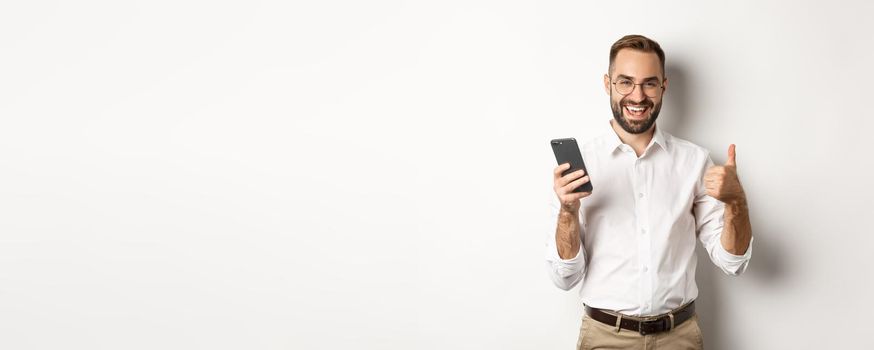 Satisfied business man showing thumbs up after using mobile phone, standing pleased over white background