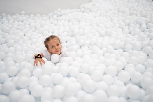 Happy little girl playing white plastic balls pool in amusement park. playground for kids.