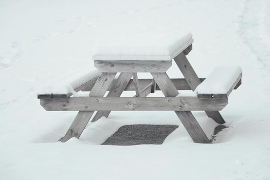 An empty bench and table in the park are covered with snow.