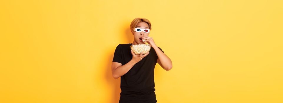 Impressed and thrilled asian guy attend cinema, watching movie or tv series in 3d glasses, eating popcorn and looking shocked, standing yellow background
