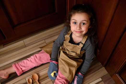 Top view cute child girl, little baker confectioner in a beige chef apron and kitchen mittens sweetly smiling to camera