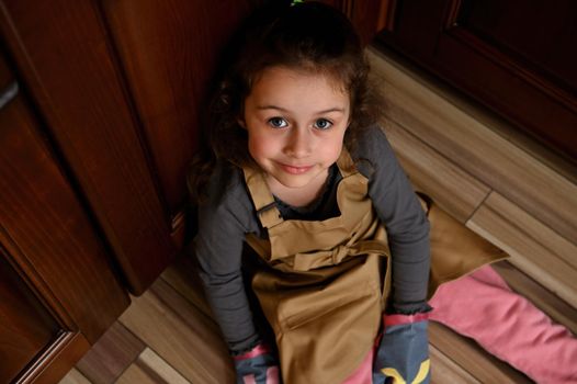 Top view of little child girl, baker confectioner in chef apron sitting barefoot on kitchen floor and looking at camera