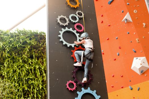 Child climbing on wall in amusement centre. Climbing training for children. Little girl in dressed climbing gear climb high. Extreme active leisure for kids.