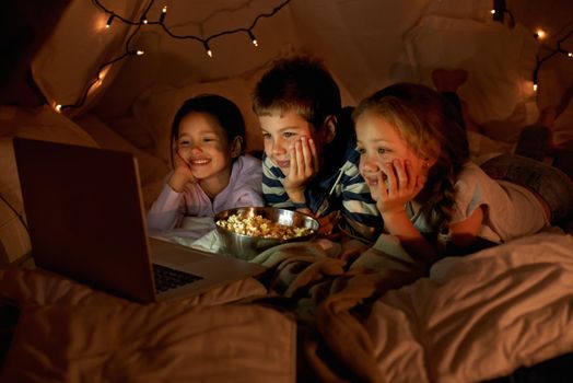 Movie time in the pillow fort. three young children using a laptop in a blanket fort.