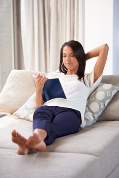Hooked on a book. an attractive young woman reading a book while reclining on the sofa.