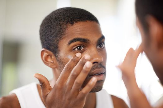 He takes care of his skin. A young man applying cream to his face while looking in the mirror.