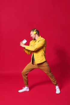 Full portrait of singing handsome man enjoying his favorite song using phone and wireless headphones wearing casual jacket isolated on red background. Man sing while recording his voice