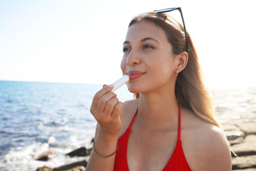 Smiling young woman applying sun protection on her lips on the beach