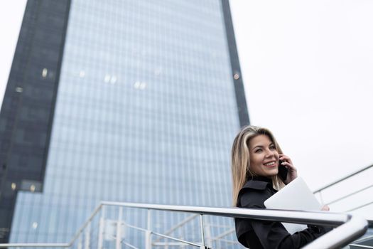 Business woman blonde against the backdrop of an office skyscraper expired with a mobile phone and a laptop